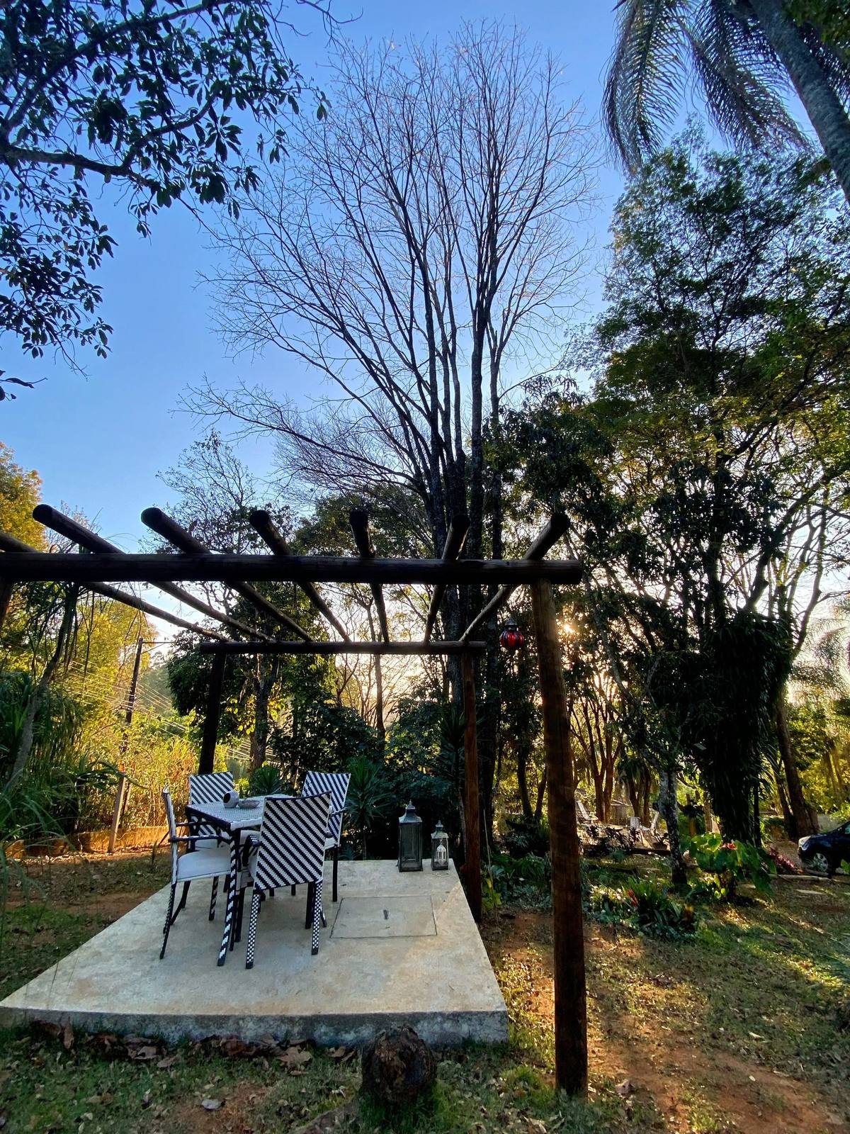 Retáculo de pérgola de madeira sobre lajota de concreto, com mesa e cadeiras, ao entardecer, entre árvores